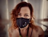A woman with curly red hair is wearing a black face mask with a filter. Her eyes are expressive and she is looking directly at the camera. The background is blurred, focusing the attention on her face.