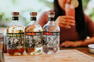 Three bottles of Espolòn tequila are displayed on a wooden surface. Each bottle has a distinct label featuring black and white illustrations. In the background, a person is holding a pink cocktail garnished with a lime slice.