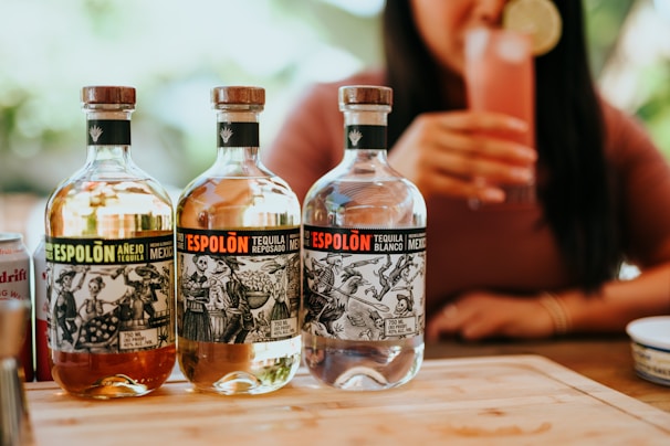 Three bottles of Espolòn tequila are displayed on a wooden surface. Each bottle has a distinct label featuring black and white illustrations. In the background, a person is holding a pink cocktail garnished with a lime slice.