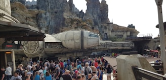 A large crowd of people gather in front of a massive, detailed spacecraft resembling a famous fictional ship. The backdrop consists of rocky, mountainous structures and what appears to be a themed amusement park or attraction. The setting is lively, with numerous visitors exploring the area.