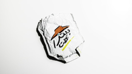 A crumpled piece of white paper featuring Arabic text and a logo with a brown hat. The paper is set against a plain white background, showing wrinkles and irregular shapes due to its crumpled state.