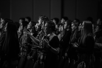 A group of people standing closely together in a dimly lit environment, appearing focused and engaged. Their expressions suggest attentiveness, and some have their hands raised, possibly indicating a moment of concentration or worship. The scene is captured in black and white.