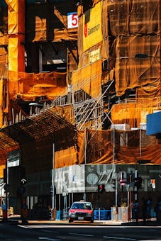 A construction site with scaffolding and orange netting covering a building, which is partially under construction. A taxi is parked in front of the site, and there are traffic lights and pedestrians visible. Signage and safety barriers are present around the area.