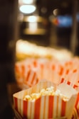 Several popcorn bags with red and white stripes are filled with popcorn, set against a softly focused, dimly lit background, suggesting a cinema or concession stand setting.