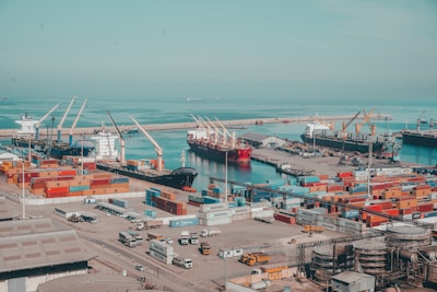 A bustling commercial port with multiple cargo ships docked at the harbor. Numerous colorful shipping containers are stacked in various configurations. Cranes are actively positioned around the dock, indicating an operational environment. The sea in the background is calm, and there are some smaller ships visible in the distance.