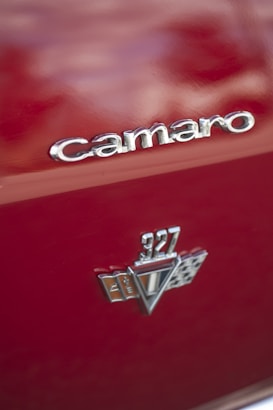 Close-up of a red car body featuring the 'Camaro' badge and a '327' emblem with racing flag details, which are both in chrome.