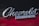 Chrome 'Chevrolet Camaro' emblem mounted on the exterior of a car. The background features a glossy surface with a red and black color scheme.