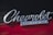Chrome 'Chevrolet Camaro' emblem mounted on the exterior of a car. The background features a glossy surface with a red and black color scheme.