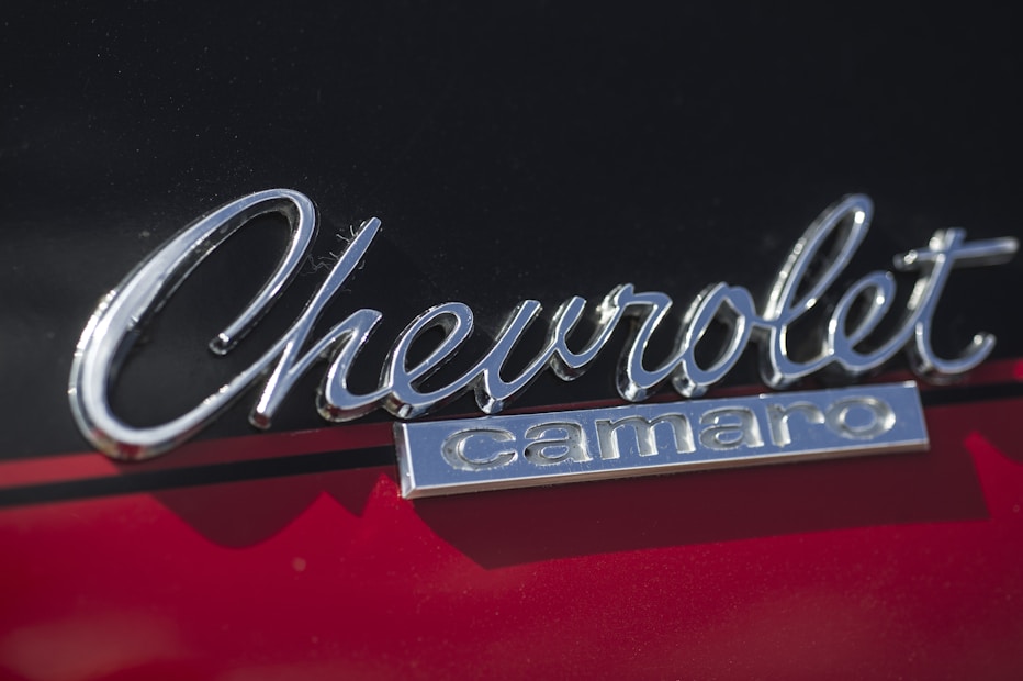 Chrome 'Chevrolet Camaro' emblem mounted on the exterior of a car. The background features a glossy surface with a red and black color scheme.