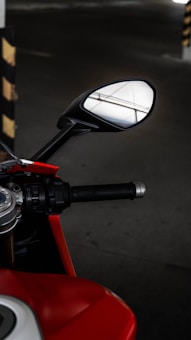 A close-up view of a motorcycle's handlebar and side mirror, showcasing detailed parts like the grip, control buttons, and a glimpse of the fuel tank. The mirror reflects a portion of a ceiling with structural lines. The scene is set in a dimly lit space, likely a parking area, with a black and yellow striped pole visible in the background.