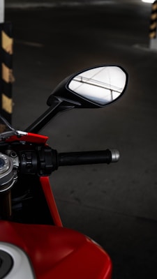 A close-up view of a motorcycle's handlebar and side mirror, showcasing detailed parts like the grip, control buttons, and a glimpse of the fuel tank. The mirror reflects a portion of a ceiling with structural lines. The scene is set in a dimly lit space, likely a parking area, with a black and yellow striped pole visible in the background.