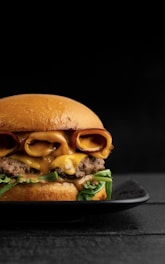 A gourmet hamburger is placed on a black plate, featuring layers of melted cheese, crispy bacon, fresh green lettuce, and a succulent beef patty, all encased in a glossy brioche bun. The background is dark, highlighting the appetizing colors and textures of the burger.