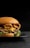 A gourmet hamburger is placed on a black plate, featuring layers of melted cheese, crispy bacon, fresh green lettuce, and a succulent beef patty, all encased in a glossy brioche bun. The background is dark, highlighting the appetizing colors and textures of the burger.