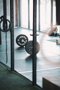 A gym setting with a barbell loaded with weights placed on the floor. The background shows gym equipment such as rings and possibly weightlifting platforms. Lighting appears to be natural, coming through large windows.