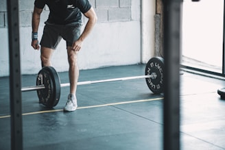 A person is dressed in athletic wear, adjusting weights on a barbell in a gym setting. The floor is marked with lines and the environment is minimalistic with concrete walls.
