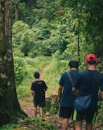 Three people are walking along a lush, green forest trail. The path is narrow and surrounded by dense foliage and tall trees. One person wears a red hat with white spots and has a blue sling bag, while the others are dressed in casual dark attire. The setting feels calm and peaceful, suggestive of a leisurely hike or nature walk.