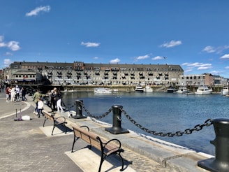 A waterfront scene featuring a row of buildings along the edge of the water. Several boats are docked in the marina, and a group of people are gathered along the promenade. A few benches face the water, and the promenade is bordered by a black chain fence with large, sturdy posts. The sky is clear with a few scattered clouds.