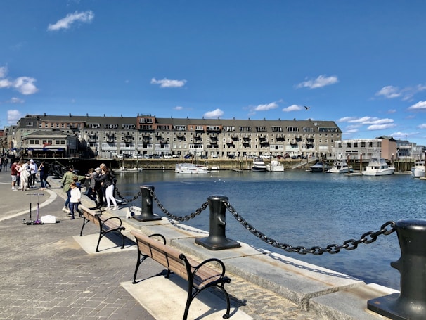 A waterfront scene featuring a row of buildings along the edge of the water. Several boats are docked in the marina, and a group of people are gathered along the promenade. A few benches face the water, and the promenade is bordered by a black chain fence with large, sturdy posts. The sky is clear with a few scattered clouds.