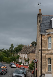 A residential street scene featuring a row of attached stone houses with chimneys and a slate roof. The street is lined with parked cars and bordered by dense greenery and trees. A 20 mph speed limit sign is visible, and there are orange construction barriers on the sidewalk.