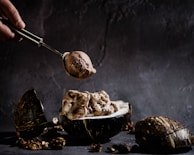 A hand holding a metal scoop is serving chocolate ice cream into a halved coconut shell. The setting has a rustic and dark aesthetic with scattered nuts around. The background is textured and adds to the moody ambiance.