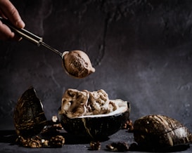 A hand holding a metal scoop is serving chocolate ice cream into a halved coconut shell. The setting has a rustic and dark aesthetic with scattered nuts around. The background is textured and adds to the moody ambiance.