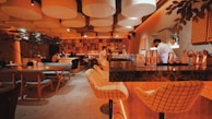A warmly lit, modern bar with a cozy atmosphere, featuring grid-patterned chairs along a dark marble counter. There are people sitting and a bartender at work. The ceiling is adorned with large, circular acoustic panels, and the walls display various framed images. Ambient lighting enhances the relaxed mood, with some greenery adding to the decor.