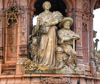 A detailed sculpture of a man and woman made from stone. The woman is standing, holding a bunch of wheat, while a sheep stands beside her. The man, dressed as a farmer, is wearing a hat and seated with his arms crossed. Both figures are part of a larger, ornately carved stone structure with intricate designs and columns. The base of the sculpture has the word 'Australia' engraved on it.