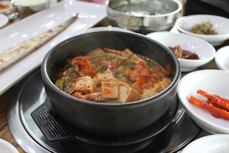 A steaming bowl of spicy stew with tofu, vegetables, and meat, served in a black stone pot. Surrounding the pot are various side dishes in white bowls, including pickled vegetables and possibly a long dish with a fish or flatbread.