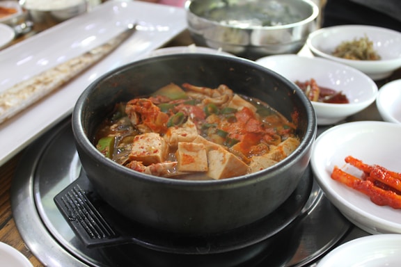A steaming bowl of spicy stew with tofu, vegetables, and meat, served in a black stone pot. Surrounding the pot are various side dishes in white bowls, including pickled vegetables and possibly a long dish with a fish or flatbread.