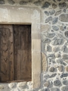 A rustic wooden shutter is set within a stone wall. The wall is constructed with a variety of irregularly shaped stones, giving a rugged and textured appearance. The wooden shutter is made of vertical planks with visible wood grain and knots. The stone wall has a beige and gray color palette, enhancing the earthy and natural look.