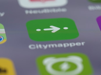 A close-up view of a smartphone screen displaying the Citymapper app icon, which is green with a white arrow and two dots. The background shows blurred app icons in various colors, creating a soft-focus effect.
