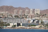 A coastal cityscape with a variety of buildings, including modern high-rise apartments and resorts, set against a backdrop of barren rocky mountains. The foreground features a beach with palm trees, sunshades, and people enjoying the waterfront.