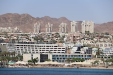 A coastal cityscape with a variety of buildings, including modern high-rise apartments and resorts, set against a backdrop of barren rocky mountains. The foreground features a beach with palm trees, sunshades, and people enjoying the waterfront.