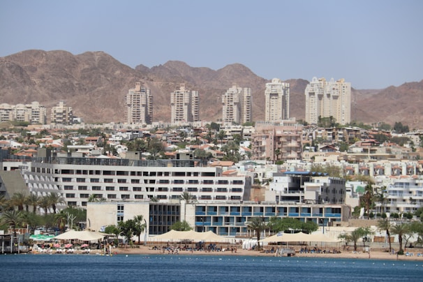 A coastal cityscape with a variety of buildings, including modern high-rise apartments and resorts, set against a backdrop of barren rocky mountains. The foreground features a beach with palm trees, sunshades, and people enjoying the waterfront.