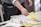 A person is organizing colorful sticky notes on a whiteboard or large sheet of paper. The person is holding a pen and appears to be working in a collaborative or planning setting. On the table, there are various objects including a coffee cup, a roll of tape, a smartphone, and some documents.