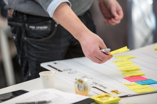 A person is organizing colorful sticky notes on a whiteboard or large sheet of paper. The person is holding a pen and appears to be working in a collaborative or planning setting. On the table, there are various objects including a coffee cup, a roll of tape, a smartphone, and some documents.