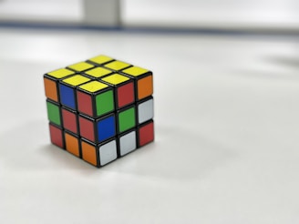 A Rubik's Cube resting on a white surface, displaying a variety of colors including yellow, red, blue, green, orange, and white. It appears well-lit, standing out against the blurred background.
