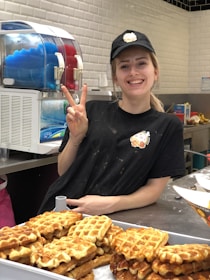 A smiling person is standing behind a counter displaying a large tray of waffles. They are wearing a black cap and shirt, both featuring a logo. In the background, there is a slushy machine with blue and red beverages.