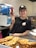 A smiling person is standing behind a counter displaying a large tray of waffles. They are wearing a black cap and shirt, both featuring a logo. In the background, there is a slushy machine with blue and red beverages.