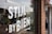A storefront window displays white block letters with the words 'STIJL BY BIEN' along with 'EST 2019'. There are reflective surfaces showing a street scene with parked cars and some greenery inside the store. The window also features hanging ornaments, and bricks are visible on the wall beside the window.