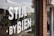 A storefront window displays white block letters with the words 'STIJL BY BIEN' along with 'EST 2019'. There are reflective surfaces showing a street scene with parked cars and some greenery inside the store. The window also features hanging ornaments, and bricks are visible on the wall beside the window.