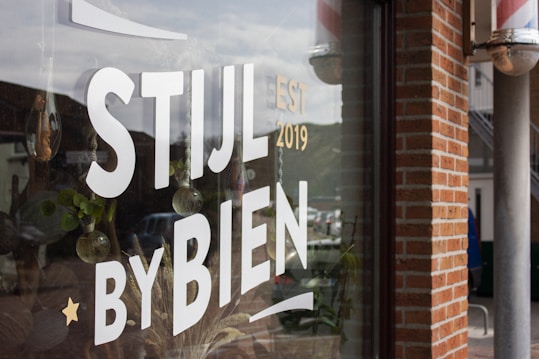 A storefront window displays white block letters with the words 'STIJL BY BIEN' along with 'EST 2019'. There are reflective surfaces showing a street scene with parked cars and some greenery inside the store. The window also features hanging ornaments, and bricks are visible on the wall beside the window.