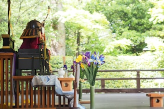 A wooden table displays traditional decorative items, including a samurai helmet with an orange and black pattern and a bow. A vase of vibrant irises in purple, yellow, and blue stands prominently. The backdrop consists of lush green trees and foliage, creating a serene outdoor setting.