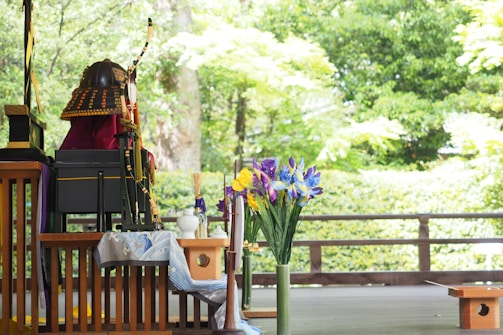A wooden table displays traditional decorative items, including a samurai helmet with an orange and black pattern and a bow. A vase of vibrant irises in purple, yellow, and blue stands prominently. The backdrop consists of lush green trees and foliage, creating a serene outdoor setting.