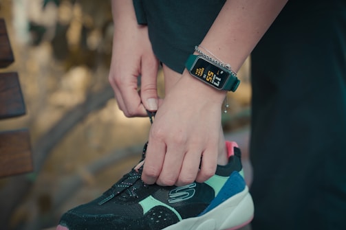 A person is tying the laces of a black, turquoise, and pink sneaker. They are wearing a digital watch that displays health tracking information on their wrist, along with a bracelet. The background is blurred, suggesting an outdoor setting.