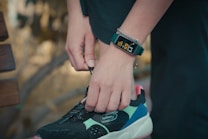 A person is tying the laces of a black, turquoise, and pink sneaker. They are wearing a digital watch that displays health tracking information on their wrist, along with a bracelet. The background is blurred, suggesting an outdoor setting.