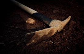 A weathered pickaxe lies on the ground, half-buried in dark, rich soil. The wooden handle is partially wrapped in a weathered grip, while the metal head is covered with dirt, indicating recent use in digging or mining. Small rocks and soil debris surround the tool, set against a dimly lit and somewhat dramatic backdrop.