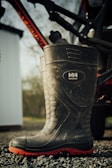 A large, rugged boot with a high shaft, designed for work purposes, is resting on a gravel surface. The boot features a visible brand logo and has a black body with a distinctive red sole. Partially visible in the background is a bicycle frame mounted on a vehicle, with a blurred outdoor scene and the edge of a white structure.