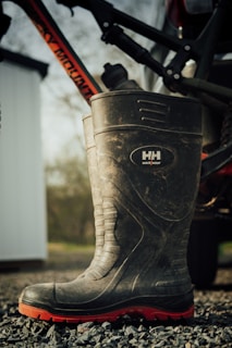 A large, rugged boot with a high shaft, designed for work purposes, is resting on a gravel surface. The boot features a visible brand logo and has a black body with a distinctive red sole. Partially visible in the background is a bicycle frame mounted on a vehicle, with a blurred outdoor scene and the edge of a white structure.