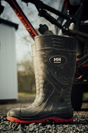 A large, rugged boot with a high shaft, designed for work purposes, is resting on a gravel surface. The boot features a visible brand logo and has a black body with a distinctive red sole. Partially visible in the background is a bicycle frame mounted on a vehicle, with a blurred outdoor scene and the edge of a white structure.